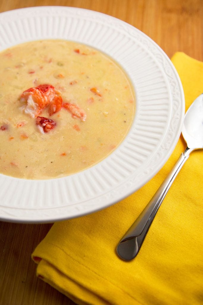 lobster chowder with lobster meat on top in a white bowl, next to a lemon yellow napkin with a spoon 