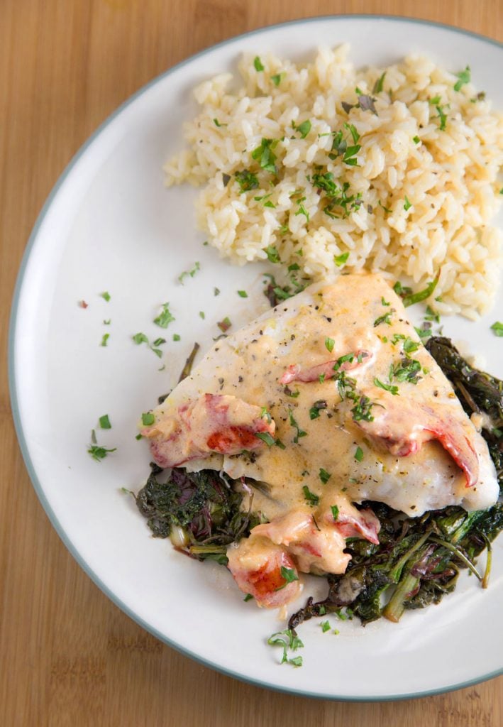 Red Snapper on a bed of greens topped with a lobster cream sauce together with rice on a white plate sitting on a cutting board