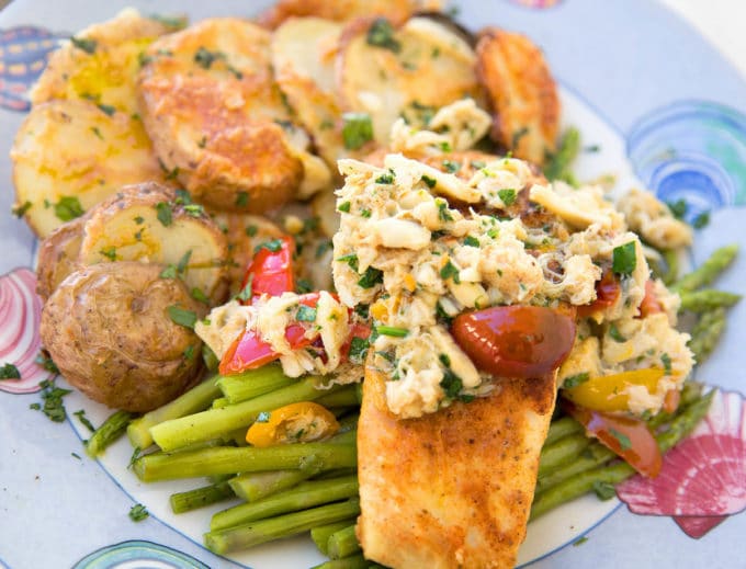 pan seared halibut with crab meat and grape tomato halves on a bed of asparagus with roasted potatoes with melted cheddar as the side dish all sitting on a blue seashore style plate 