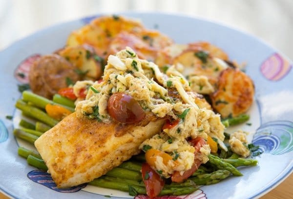 pan seared halibut with crab meat and grape tomato halves on a bed of asparagus with roasted potatoes with melted cheddar as the side dish all sitting on a blue seashore style plate 