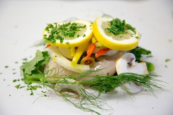 side view of Red Snapper topped with vegetables, herbs and lemon slices on a white paper