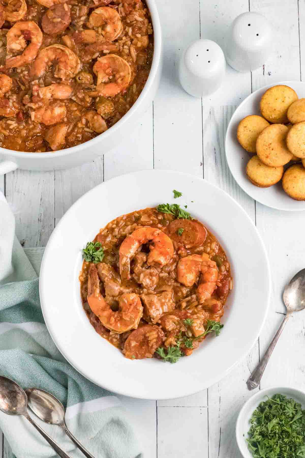 jambalaya in a white bowl with corn muffins and pot of jambalaya in background