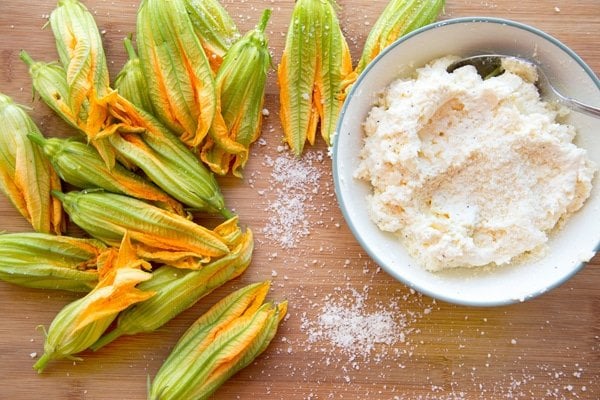 zucchini blossoms and a bowl of seasoned ricotta cheese on a wooden cutting board sprinkled with grated romano cheese