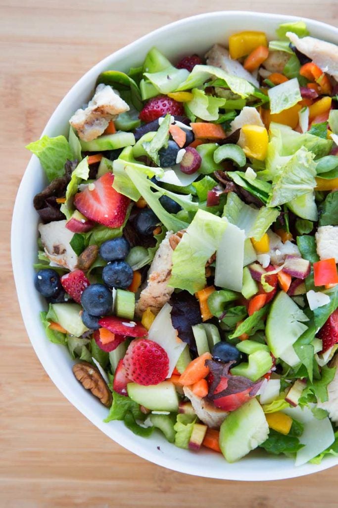 partial overhead view of a mixed summertime salad with berries, vegetables, nuts, chicken and cheese in a white bowl