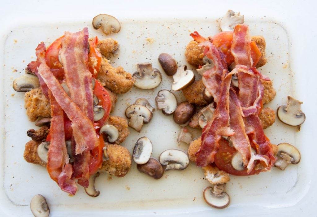 chicken strips topped with tomatoes and bacon in a baking dish with cooked sliced mushrooms