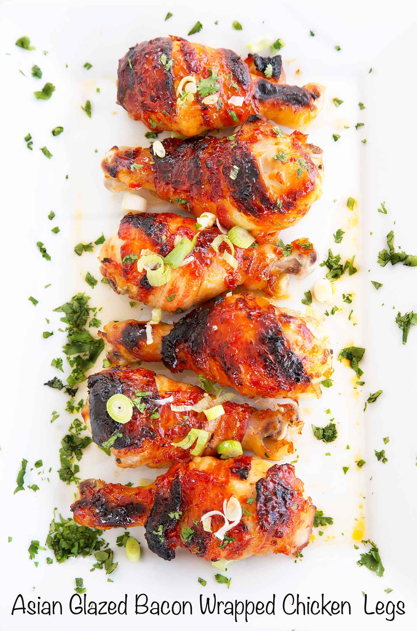 asian glazed bacon wrapped chicken legs on a white platter with sliced scallions and chopped cilantro