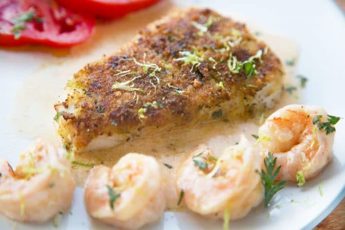 side  view of Crusted Chilean Sea Bass with a row of shrimp and the edge of tomato slices with a light sauce on a white plate