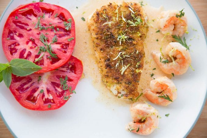 overhead view of Crusted Chilean Sea Bass with a row of shrimp and tomato slices with a light sauce on a white plate