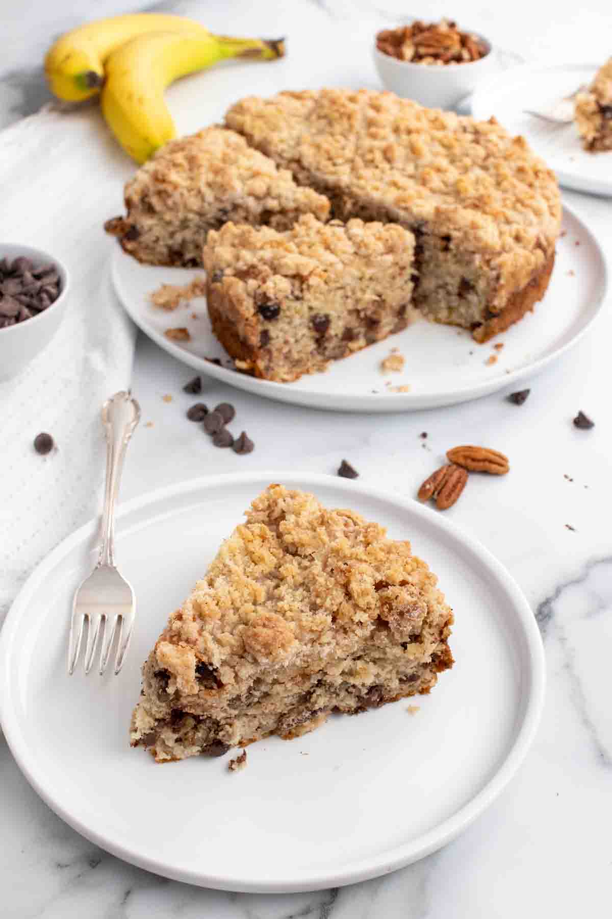 slice of banana crumb cake on a white plate with remainder of the cake in the background