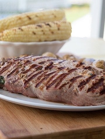 grilled stuffed pork tenderloin on a white plate on a cutting board with a bowl of grilled corn on the cob in the background