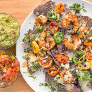 blackened shrimp nachos with melted cheese and peppers on a white plate with bowls of salsa and guacamole sitting on a cutting board