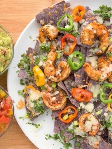 blackened shrimp nachos with melted cheese and peppers on a white plate with bowls of salsa and guacamole sitting on a cutting board