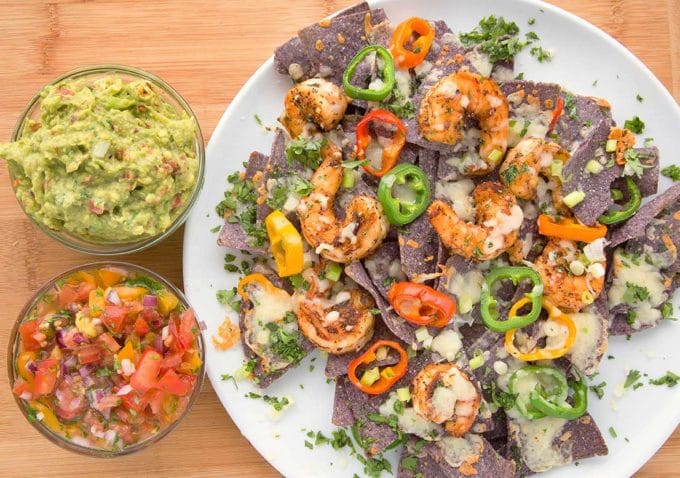 blackened shrimp nachos with melted cheese and peppers on a white plate with bowls of salsa and guacamole sitting on a cutting board