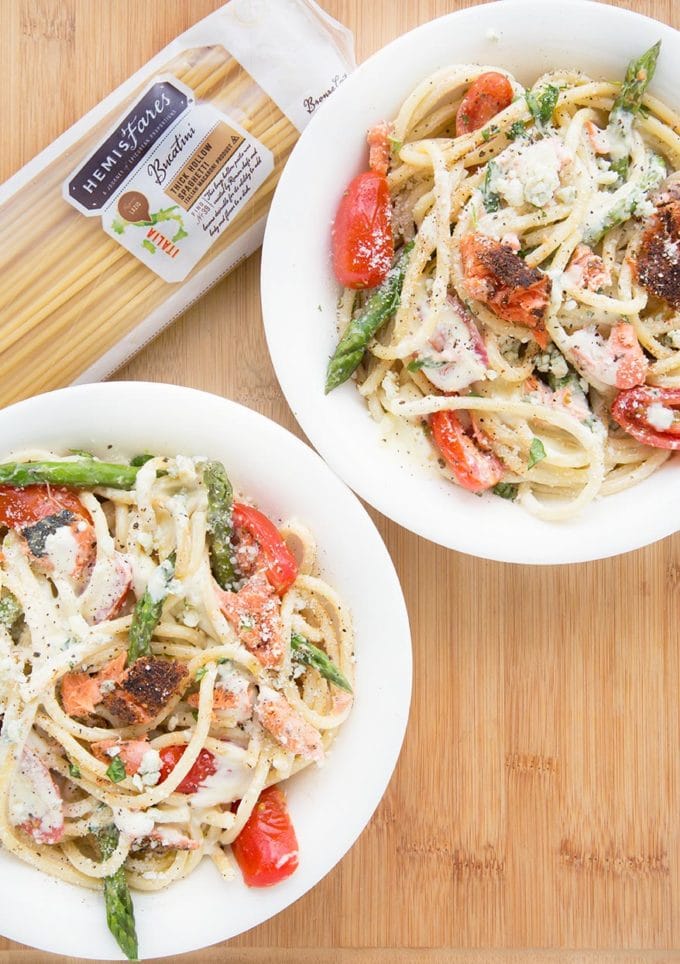 two white bowls filled with pasta mixed with salmon, asparagus, grape tomatoes and a cream sauce. sitting on a cutting board with a package of pasta