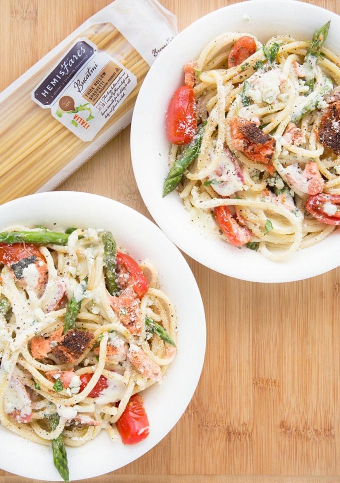 two white bowls filled with pasta mixed with salmon, asparagus, grape tomatoes and a cream sauce. sitting on a cutting board with a package of pasta 