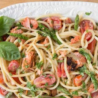 oval serving bowl filled with pasta mixed with salmon, asparagus, grape tomatoes and a cream sauce. with a sprig of basil in the corner