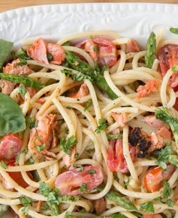 oval serving bowl filled with pasta mixed with salmon, asparagus, grape tomatoes and a cream sauce. with a sprig of basil in the corner