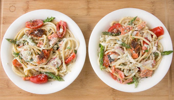 two white bowls filled with pasta mixed with salmon, asparagus, grape tomatoes and a cream sauce. sitting on a cutting board 