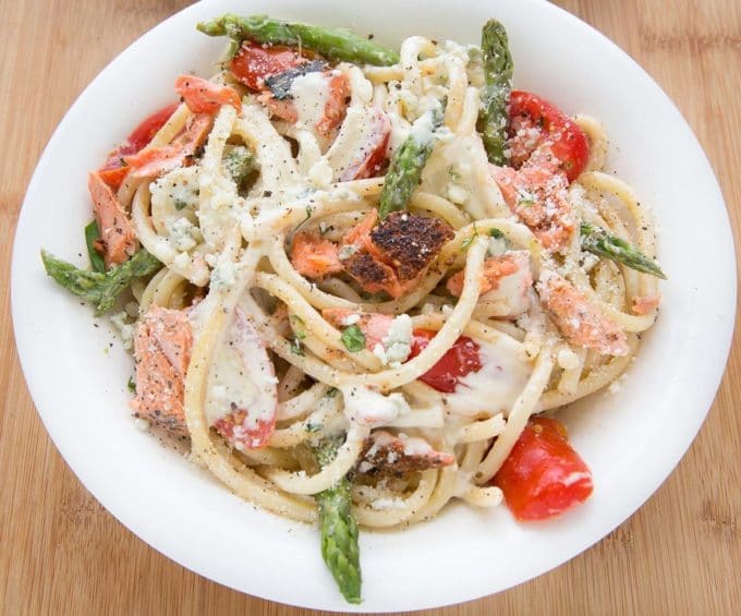 white bowl filled with pasta mixed with salmon, asparagus, grape tomatoes and a cream sauce. sitting on a cutting board