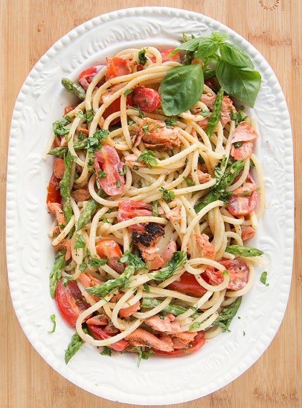 white oval serving bowl filled with pasta mixed with salmon, asparagus, grape tomatoes and a cream sauce. with a sprig of basil in the corner