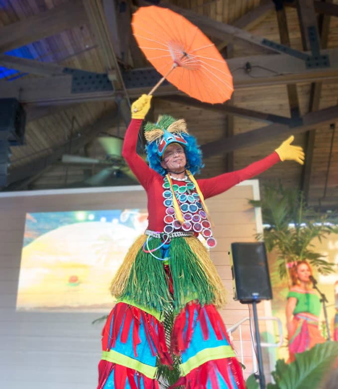 colorfully costumed man on stilts with an orange umbrella