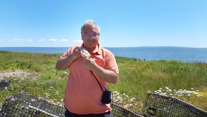 Chef Dennis holding a lobster roll in front of lobster traps with a view of the bay of fundy