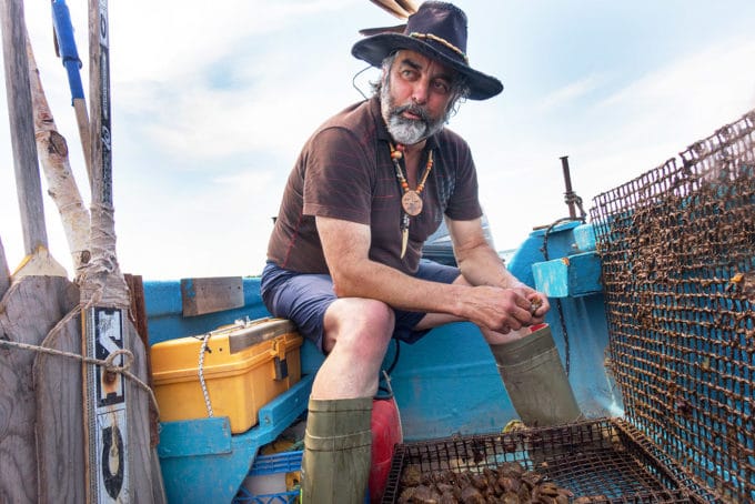 Mixed blood Acadian Oyster fisherman wearing a hat with a feather on his boat holding live oysters