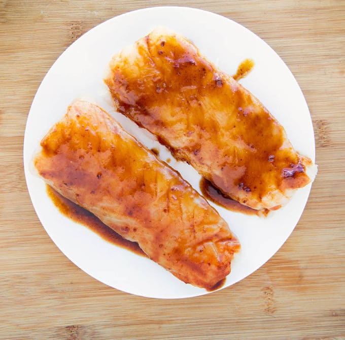 2 uncooked miso glazed cod fillets on a white plate on a cutting board