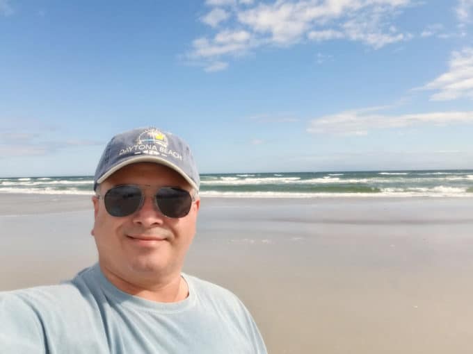 Chef Dennis at Daytona Beach in front of the ocean wearing a Daytona beach ball cap and sunglasses