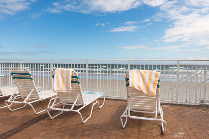 view of the ocean from a deck looking out over beach chairs with beach towels draped over them to the perfect day with blue skies and fluffy white clouds