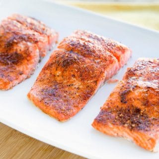 three seasoned and cooked salmon fillets on a white plate sitting on a cutting board