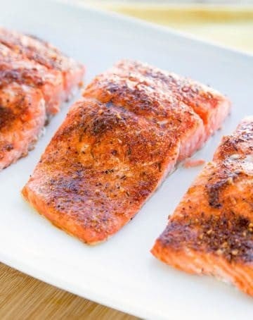 three seasoned and cooked salmon fillets on a white plate sitting on a cutting board