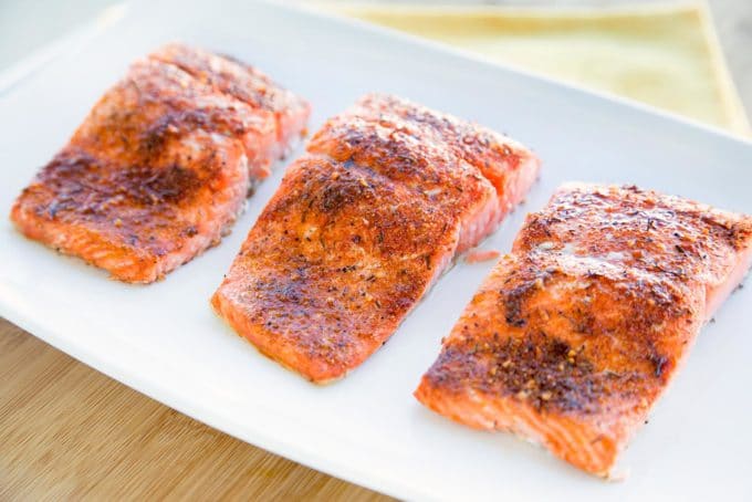 three seasoned and cooked salmon fillets on a white plate sitting on a cutting board