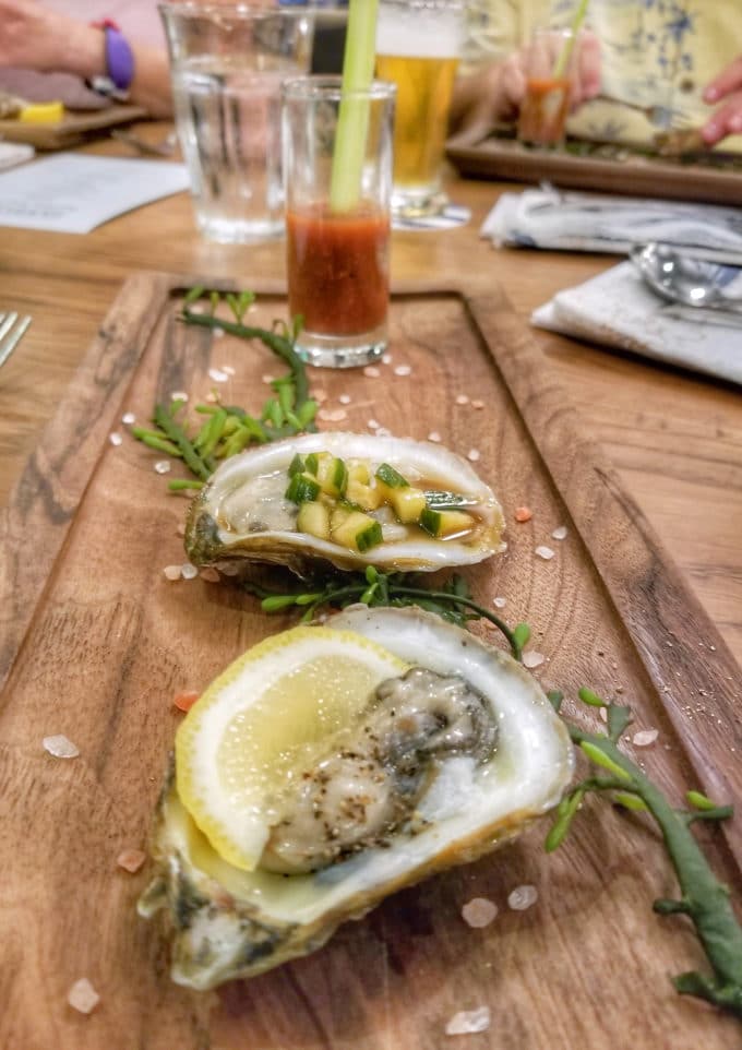 2 oysters on a wooden plank with greens around them and a shooter glass of tomato and and oyster at the end