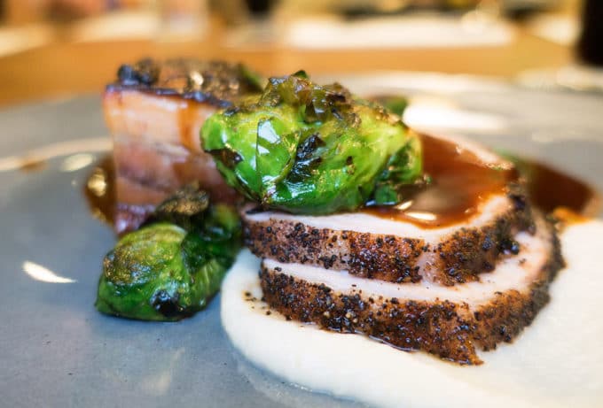 sliced pork loin, and pork belly on top of a pureed cauliflower with seared brussels sprouts and a coffee demi glace over the sliced pork and on the grey plate.