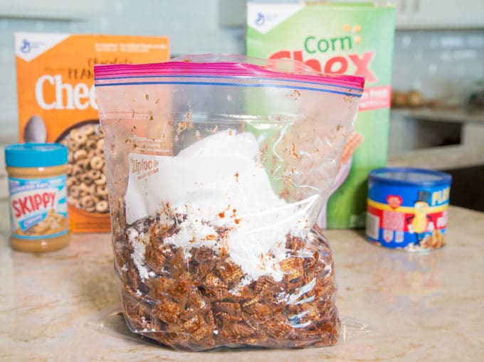 Ziplock bag full of chocolate coated chex cereal and confectioners sugar sitting on a kitchen counter with boxes of cereal in the background.