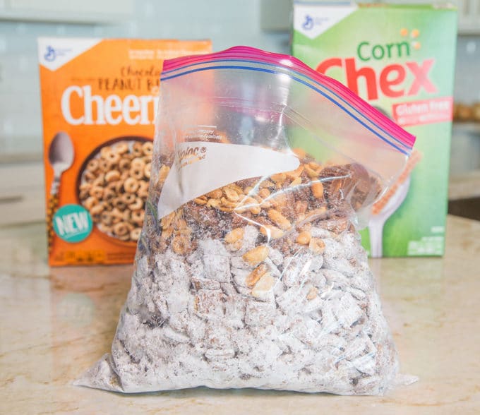 Chex cereal coated in chocolate and confectioners sugar in a ziplock bag with peanuts, peanut butter cups and cheerios sitting on a kitchen counter