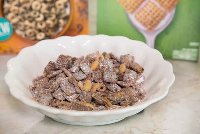 a close up shot of Chex Muddy Buddies in a white bowl with cereal boxes in the background on a kitchen counter top
