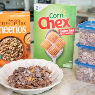 boxes of corn chex and chocolate peanut butter cheerios with Chex Muddy Buddies snack mix in a white bowl and 3 plastic ziplock containers all sitting on a counter top in the kitchen