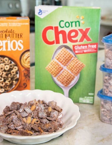 boxes of corn chex and chocolate peanut butter cheerios with Chex Muddy Buddies snack mix in a white bowl and 3 plastic ziplock containers all sitting on a counter top in the kitchen