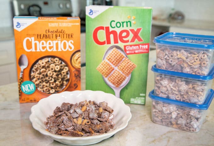 boxes of corn chex and chocolate peanut butter cheerios with Chex Muddy Buddies snack mix in a white bowl and 3 plastic ziplock containers all sitting on a counter top in the kitchen