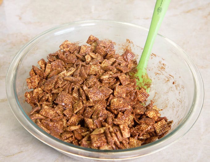 chex muddy buddies mixture in a large glass bowl with a green spatula sticking out, sitting on a kitchen counter
