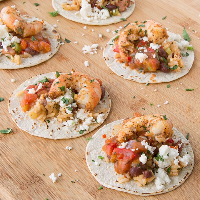 five ettouffee street tacos with a large shrimp on top all sitting on a wooden cutting board