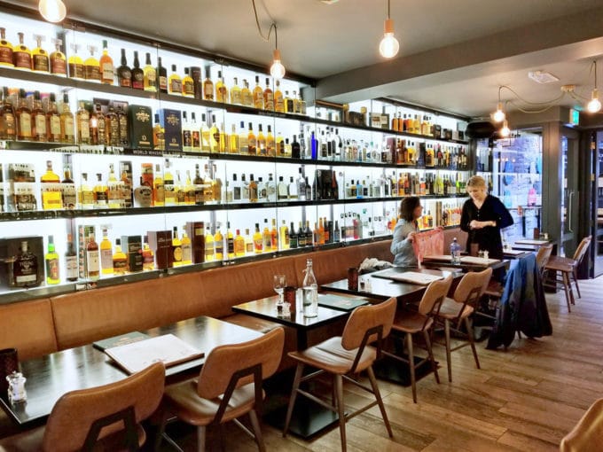 a wall of bottles of different types of alcohol with tables in front of them and woman sitting at one of the tables talking to a waitress