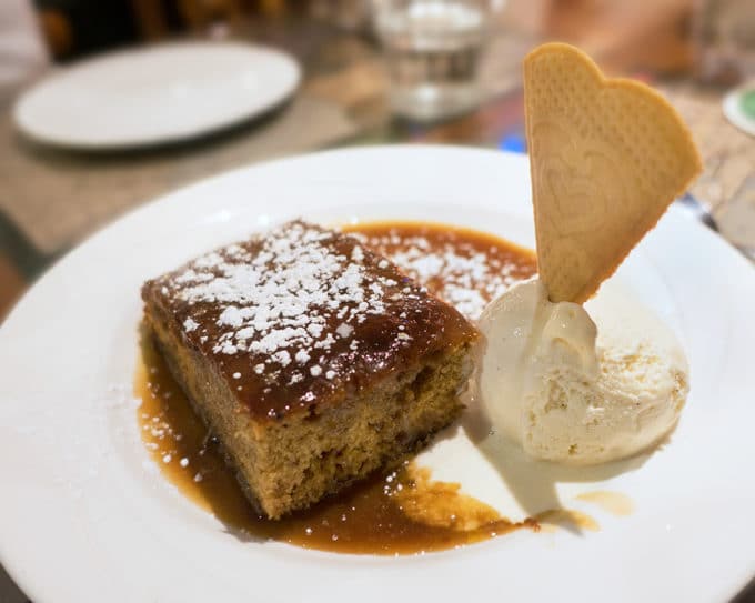 sticky toffee pudding on a white plate with whipped cream and a triangular cookie sticking out of the whipped cream