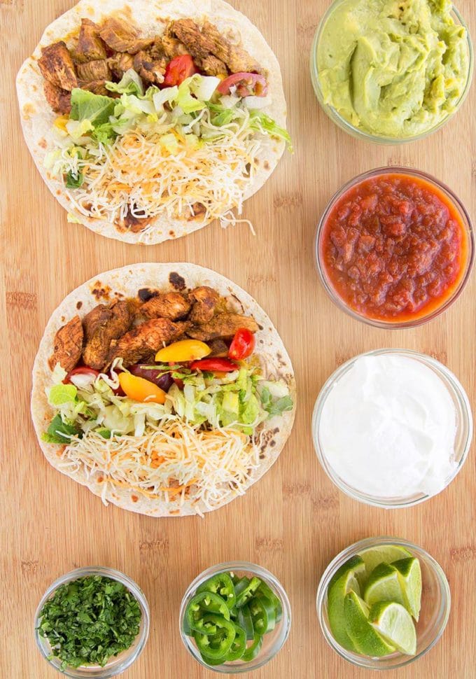 2 flour tortillas filled with blackened chicken strips, shredded lettuce sliced grape tomatoes, and Mexican blended cheese. Sitting on a wooden cutting board with bowls of guacamole, salsa, sour cream, lime wedges, jalapeno's and cilantro