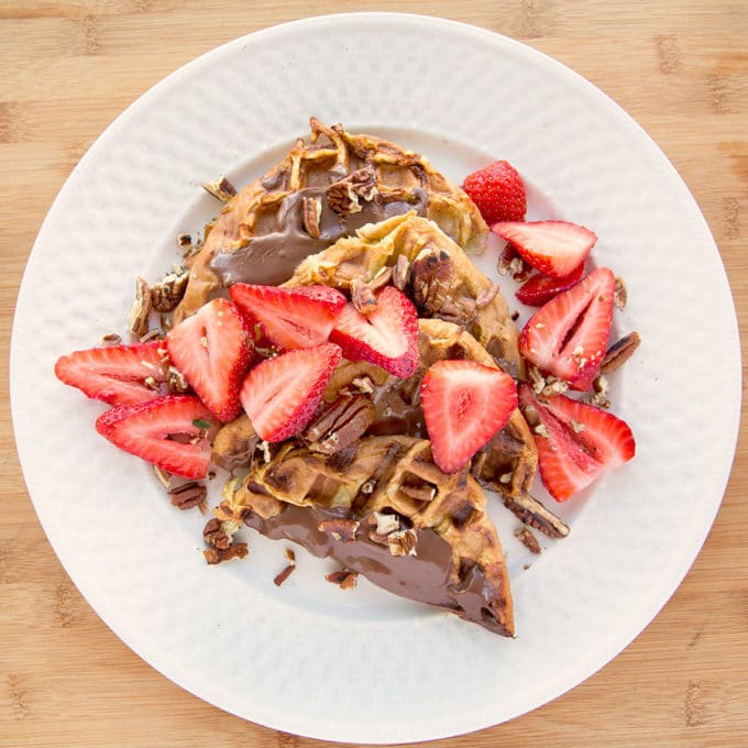 four halves of waffle iron banana stuffed french toast with nutella spread on top with sliced strawberries and toasted pecans all sitting on a white plate