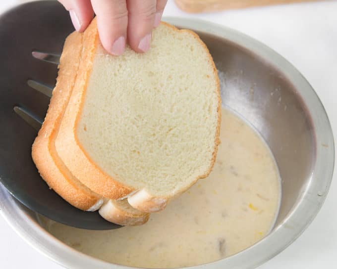 french toast being lowered into a stainless steel bowl of banana egg batter with a black spatula 