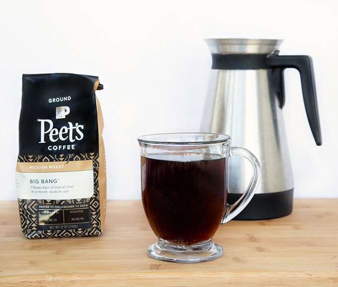 Bag of Peet's Coffee and box of Peet's Coffee K-Cups with a glass mug of black coffee sitting on a cutting board