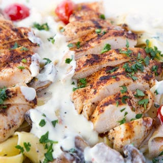 slices of seasoned grilled boneless skinless chicken breast sitting on top of Pappardelle with alfredo sauce, grilled cherry tomatoes and mushrooms sprinkled with chopped parsley